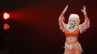 Photo of Dolly Parton in a brightly colored outfit with fringes. She's waving her hands up at a concert in 2014.