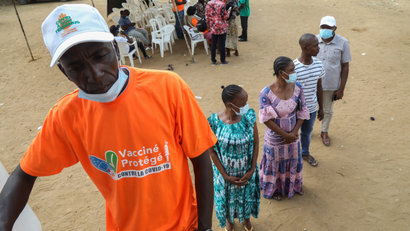 A man in an orange T-shirt and white cap stands in line. Behind him are two men and one man. Nearby are more people.