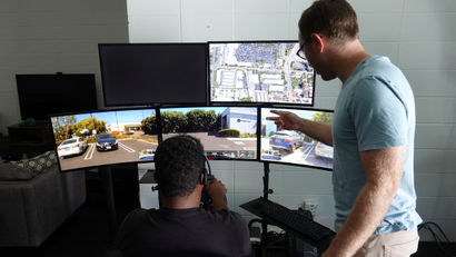 A man sits in front of five computer monitors showing different displays from a self driving car while a colleague peers over his shoulder.