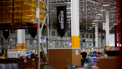 An Amazon flag flies above a collection of shelves and cardboard boxes at an Amazon warehouse.