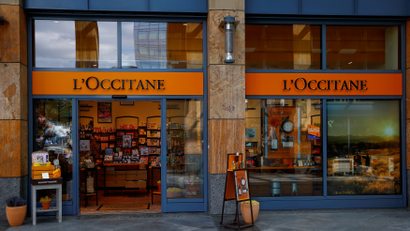 A storefront of the French beauty company L'Occitane en Provence is shown with its glass doors open and items displayed on a small table to the left.