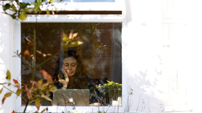 a woman sitting at her laptop, glimpsed through the window of her home