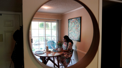 A remote worker seated before a laptop at her kitchen table is seen in a mirror's reflection