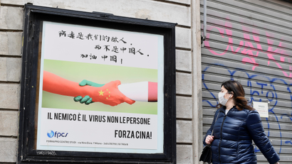 A woman, wearing a protective face mask, walks past a placard showing a handshake between two hands representing the Chinese and the Italian national flags