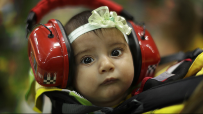 A baby girl hanging in a sling wears ear protectors