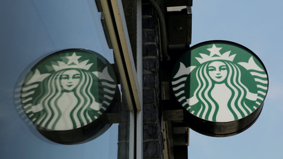 A Starbucks sign featuring its logo is reflected in the window of a store