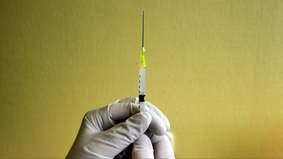 A person wearing white latex gloves holds a syringe against a yellow wall.