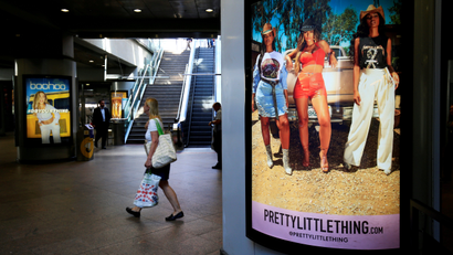 A shopper walks pass advertising billboards for Boohoo and for 'Pretty Little Things', a Boohoo brand, at Canary Wharf DLR station in central London