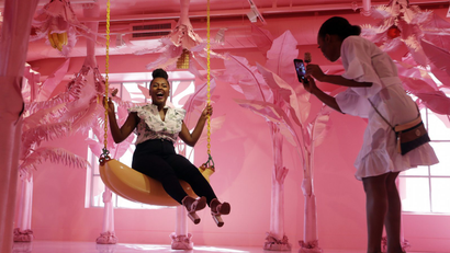 A woman sits on a banana swing while her friend takes a photo at the Museum of Ice Cream