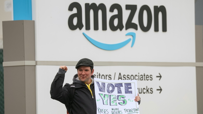 an amazon labour union organizer holding a sign that says "vote yes" stands in front of an amazon warehouse sign, with his fist raised