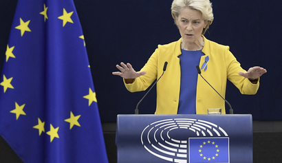 Ursula von der Leyen is standing at a podium, speaking, with both hands raised, wearing a bright yellow blazer and blue top. The podium is emblazoned with a stylized image of a round amphitheater and the EU flag. To the left of her is a large, draped EU flag.