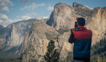 A man in a vest and baseball cap stands on the edge of a cliff taking a photo of the surrounding mountains with his iPhone.