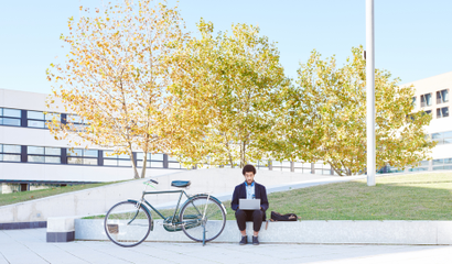 Person sitting outside working on laptop with bike next to them, trees in background
