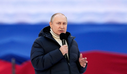 Vladimir Putin speaking into a microphone, wearing a turtleneck and a black puffer coat, standing in front of a Russian flag.