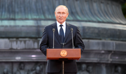 Russian president Vladimir Putin delivers a speech outside at a wooden podium. He is in front of what appears to be the base of a large, copper-oxidized monument. He wears a black suit, white shirt, and black tie with diagonal lines of white dots.