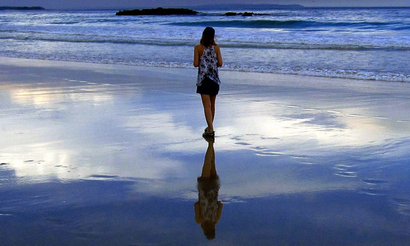 A woman walks on a beach; her reflection visible in the water