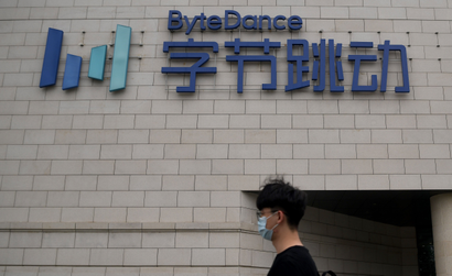 A figure of a man from the shoulders up wearing a face mask, black tshirt, and glasses can be seen in front of the cream-tiled facade of ByteDance headquarters. Above him is a lettered 3D sign with the English name "ByteDance" and Chinese name underneath in a navy blue. To the left is ByteDance's logo, which is four columns of varying lengths and heights in different shades of blue.