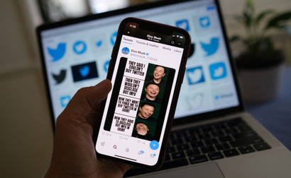 An iphone is held in someone's hand featuring a meme Musk posted to Twitter. In the background is a laptop, out of focus, displaying Twitter images.