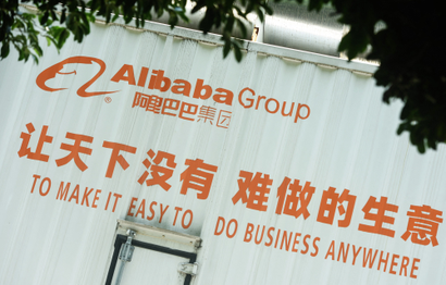 The side of what appears to be a white shipping container depicts the words "Alibaba Group" with its logo in English and Chinese, and then beneath 10 Chinese characters with the English translation "To make it easy to do business anywhere" placed beneath it. The top of the photo shows some leaves, as if the photo was taken from beneath a tree.