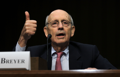 Justice Stephen Breyer sits at a desk, giving a thumbs up, captured mid-speech. There is a white name card in front of him, and a dark background.