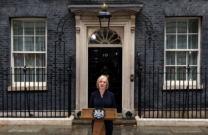 A symmetrically framed photo depicts Liz Truss standing at a podium outside the black facade of 10 Downing street, captured mid-speech. She is dressed in a dark navy dress. The pavement is wet from a rain shower. 