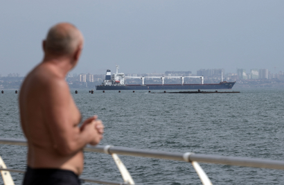 A shirtless middle-aged man stands to the left of the frame, out of focus, staring out at a cargo ship on the water. In the distant background are buildings. The sky is hazy grey-blue, and the water looks dark green.