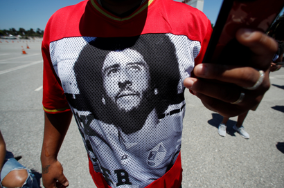 A Black man wears a t-shirt with Colin Kaepernick's face on it while holding a phone.