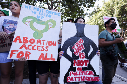 Two protesters are holding large signs, one reads "We must continue to speak up, abortion access for all," and the other sign reads, "This body is not a political battleground."