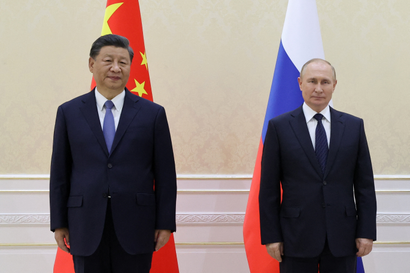 On the left is Chinese president Xi Jinping dressed in a black suit, white shirt, and periwinkle blue tie, standing in front of China's flag. Next to him stands Russian president Vladimir Putin in a black suit, white shirt, and dark navy tie with diagonal dotted stripes, standing in front of Russia's flag. The wall behind them is a buttery yellow patterned wallpaper. At the bottom is a white baseboard with three vertical, thin gold stripes.