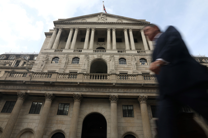 A view from below looking up at the Greco-Roman, white stone facade of the Bank of England. Entering from the right side of the frame is a man in a dark suit, appearing to be walking briskly forward. His figure is a blur. The sky above is a slightly washed out blue with wispy clouds.