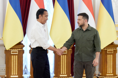 Indonesian leader Joko Widodo in a white dress shirt and black trousers shaking hands with Ukrainian president Volodymyr Zelenskyy in his trademark olive green, in front of some flags.