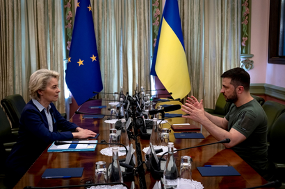 Ursula von der Leyen and Volodymyr Zelenskyy sit across from each other at a conference table, she in a suit, he in his customary olive T-shirt, and the flags of the EU and Ukraine in the background. Zelenskyy is speaking and gesturing with his hands, and von der Leyen is listening with her hands on the table.