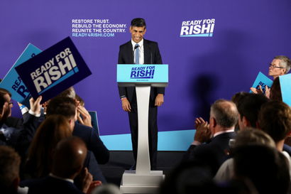 Rishi Sunak stands at a podium giving a cheeky smile. Behind him is a deep purple-blue backdrop that reads "Ready for Rishi." Supporters crowd in the foreground, raising signs with the same slogan.