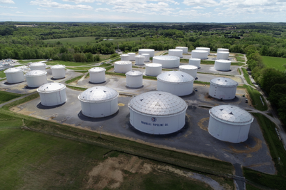 An aerial view of several large, cylindrical fuel holding tanks set against a backdrop of fields and trees.