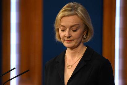 A portrait of Liz Truss, her eyes looking down while standing at a podium. She is wearing a black blazer, a gold necklace, and gold earrings.