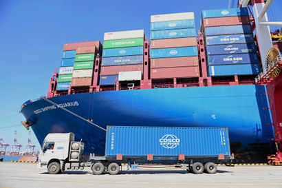 A truck drives through a port trailing a blue "Cosco" shipping container. Behind it is a blue-hulled ship stacked with shipping containers in a variety of colors. The sky above is a clear blue.