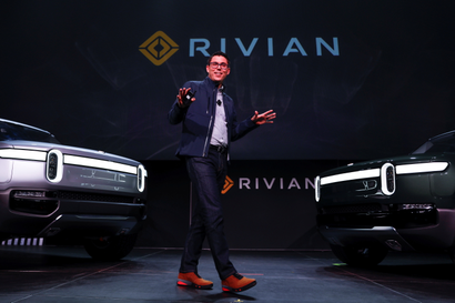 The founder of electric vehicle company Rivian stands in between two trucks with the company's logo above.