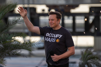 A portrait of Elon Musk with one hand raised, waving, and the other holding a mic. He is smirking slightly and wearing a black t-shirt that reads "Occupy Mars."