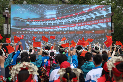 An audience sits in front of a screen displaying the large conference hall filled with delegates attending the 20th Chinese Communist Party Congress. Audience-members are raising and waving China's flag.
