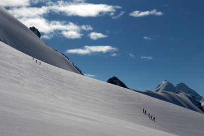 Mountain climbers tied together make their way up a steep, snowy peak with a blue sky in the background.