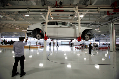 A man stands on a shiny white Tesla factory floor, holding up a phone camera to an unfinished Tesla model hanging from the ceiling. The car is white and has no tires yet.