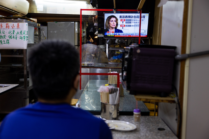 In the foreground, blurry, is a man sitting. We can see the back of his head and his blue shirt. He appears to be in a restaurant, the table in front of him has an empty plate and a container of chopsticks. Further in the background is a TV with a news broadcast in Chinese showing a photo of US House speaker Nancy Pelosi.