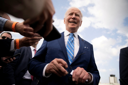 US president Joe Biden smiling in front of reporters. The photo is taken from waist-height, and you can see blue sky behind him.