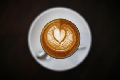 A latte in a white cup and saucer with a black background and heart in the foam on top.