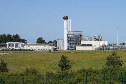 An white factory with a smokestack cuts horizontally across the center of the photo, the foreground is a grass field with shrubs. The sky in the background is blue. 