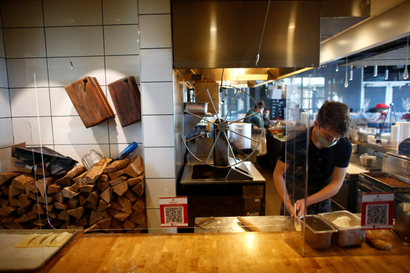 Jesse Mobley stands on the right of the photo wearing a black t-shirt and a mask while he prepares pastries in a Denver restaurant kitchen behind sheets of plexiglass. A large pile of logs is on the left of the photo against a white-tiled alcove, along with several wooden boards hanging on the wall. Four pastries sit on a parchment paper lined metal sheet on the butcher-block counter.
