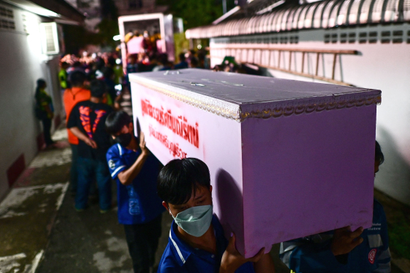 Men wearing masks and blue polo shirts carry a light pink coffin with red lettering down a street. In the background are more people, blurry, and the open back of a truck where more coffins are being lifted out.
