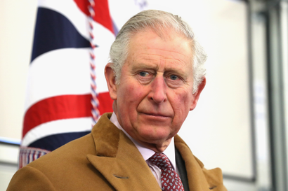 A portrait of King Charles III wearing a tan coat, over a light pink shirt, maroon tie, and what looks like a gray suit. His complexion is slightly ruddy as he looks off to the left, with the hint of a smile. The UK flag hangs in the background.