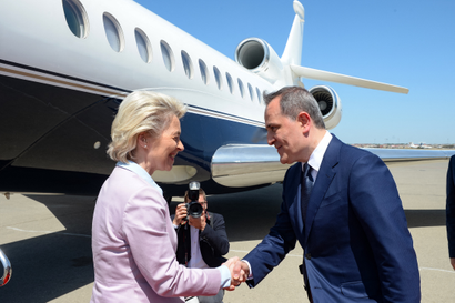 European Commission president Ursula von der Leyen and Foreign Minister of Azerbaijan Jeyhun Bayramov shake hands on the tarmac in front of a plane. Von der Leyen is dressed in a pastel pink suit, Bayramov in a dark blue suit. Both are smiling.