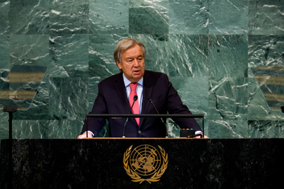 Antonio Guterres stands at a long podium made of black marble with the gold UN logo at the front, wearing a dark suit jacket, red-pink tie, and light blue shirt. Behind him is a wall of green marble tiles.